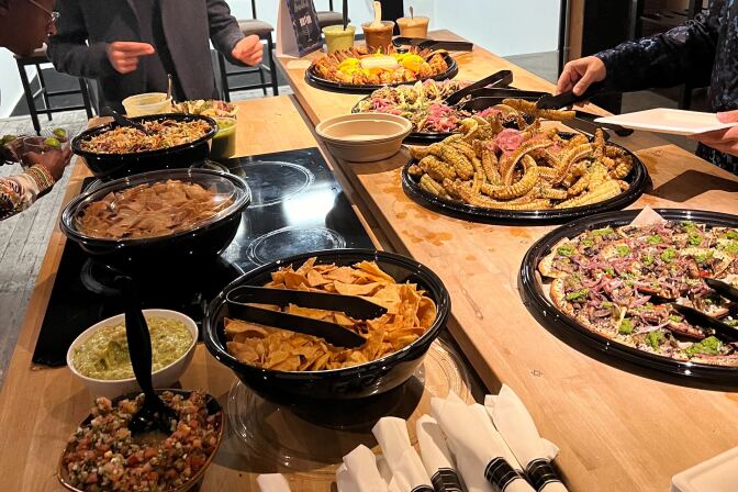 A man with facial hair wears a dark suit while bowing and gesturing over multiple plates of food at a table.