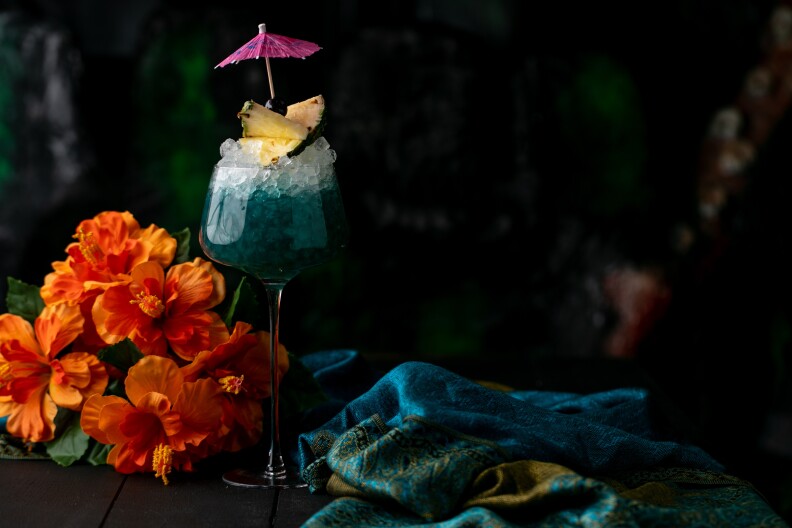 a tall wine glass filled with teal liquid and topped with a wedge of pineapple and a cocktail umbrella, in front of a bouquet of orange flowers