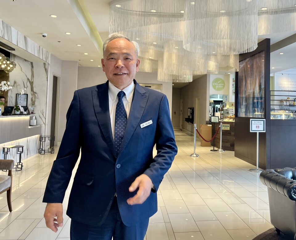 A white-haired Japanese man in a suit poses in small lobby with cream-colored marble floors. 