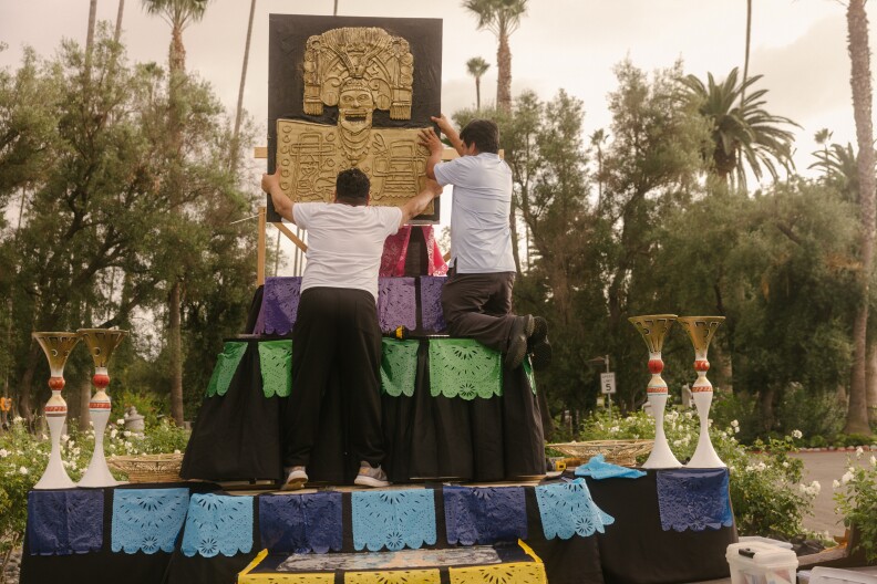 Two men, seen from behind, are working on setting up an altar in a park-like area where many trees and plants, including rose bushes, are seen in the background: The altar has several levels to it, and each level is accented with brightly colored flags. Several ornamental vases are on either side of the altar, awaiting flowers.