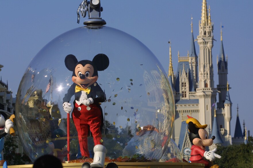 A mascot of Mickey Mouse in formal attire--a black tuxedo, yellow bow tie, white button down, and red pants--stands in a large transparent globe with golden confetti sprinkled around inside of it . Behind the mascot is the large grey, baby blue and golden Disney castle under a bright blue, cloudless sky.