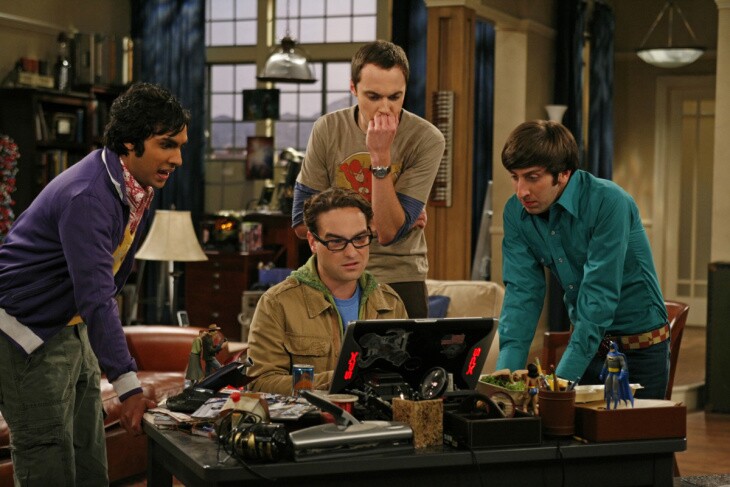 Four men gather around a desk where one man in glasses is sitting in front of a computer. Mountains are visible in the window behind
