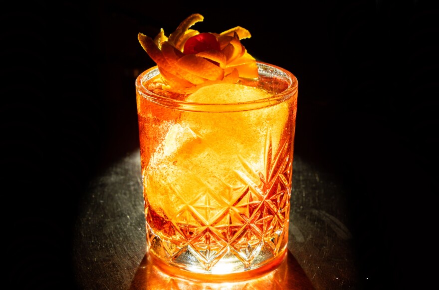 A clear glass filled with amber liquid and ice sits on a dark bar surrounded by darkness.