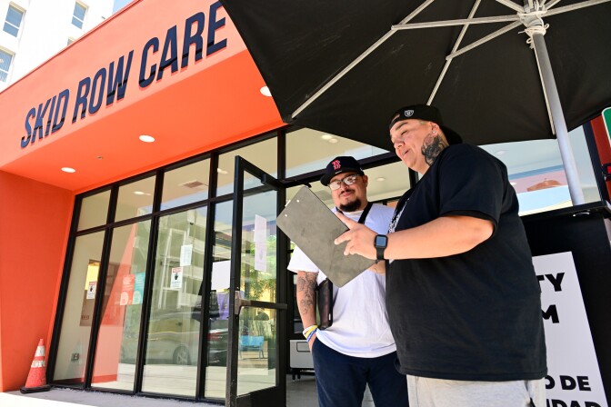 A person wearing a backwards cap and holding a clipboard stands with another person under an umbrella in front of a building which says "Skid Row Care (Campus)"