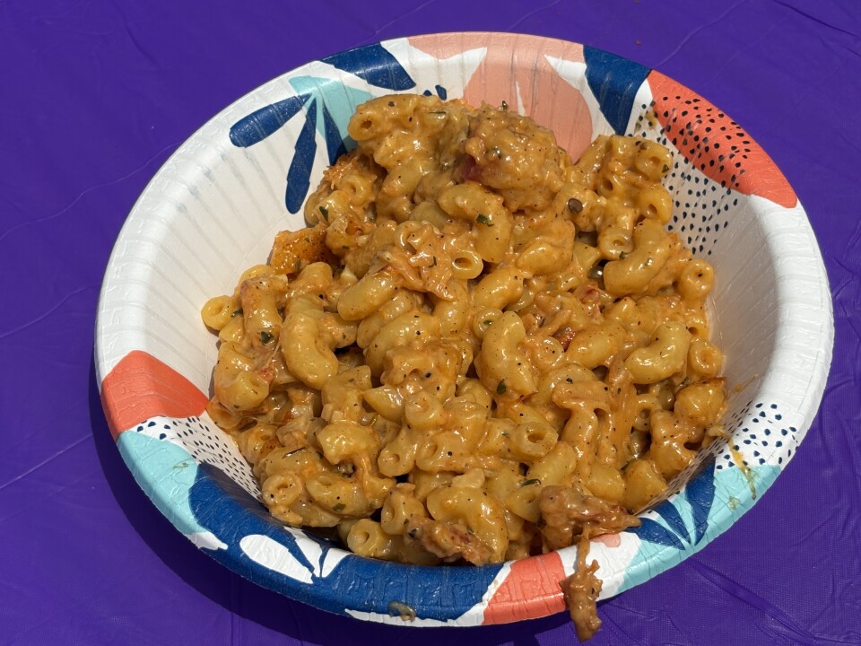 A mac and cheese coated in a thick, creamy, orange-hued cheese sauce served in a colorful, tropical-themed disposable bowl atop a vibrant purple tablecloth.