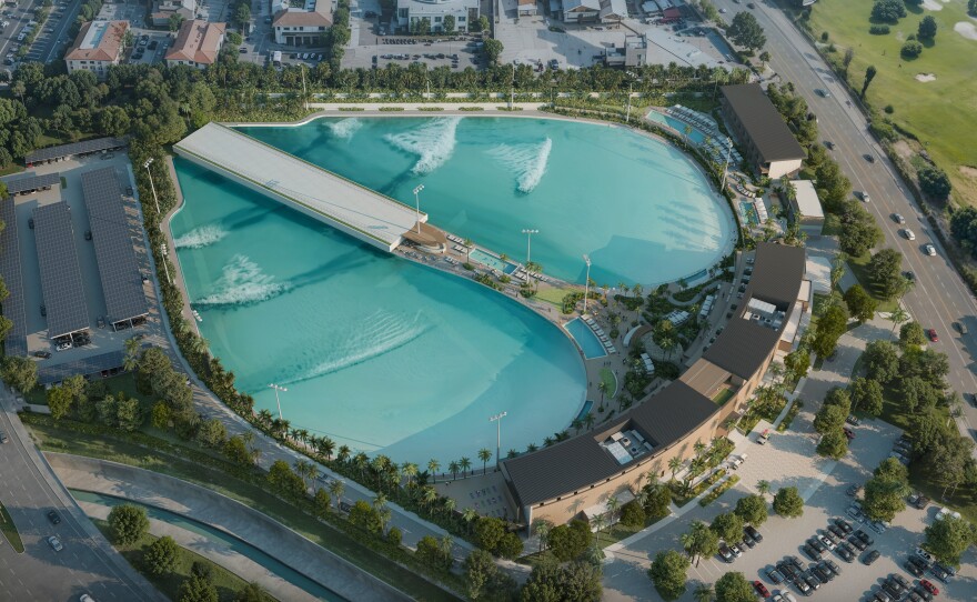 Two pools form a heart shape with a type of pier running down the middle of it: Uniform, generated waves are making their way across the surface. The exterior of the pools are lined with palm trees and building with patios where onlookers can catch the action.  