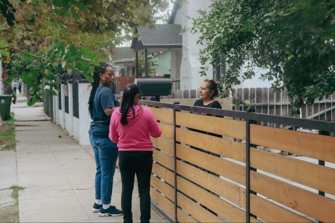 Two women stand at a wooden fence, speaking to another woman on the other side of the fence.
