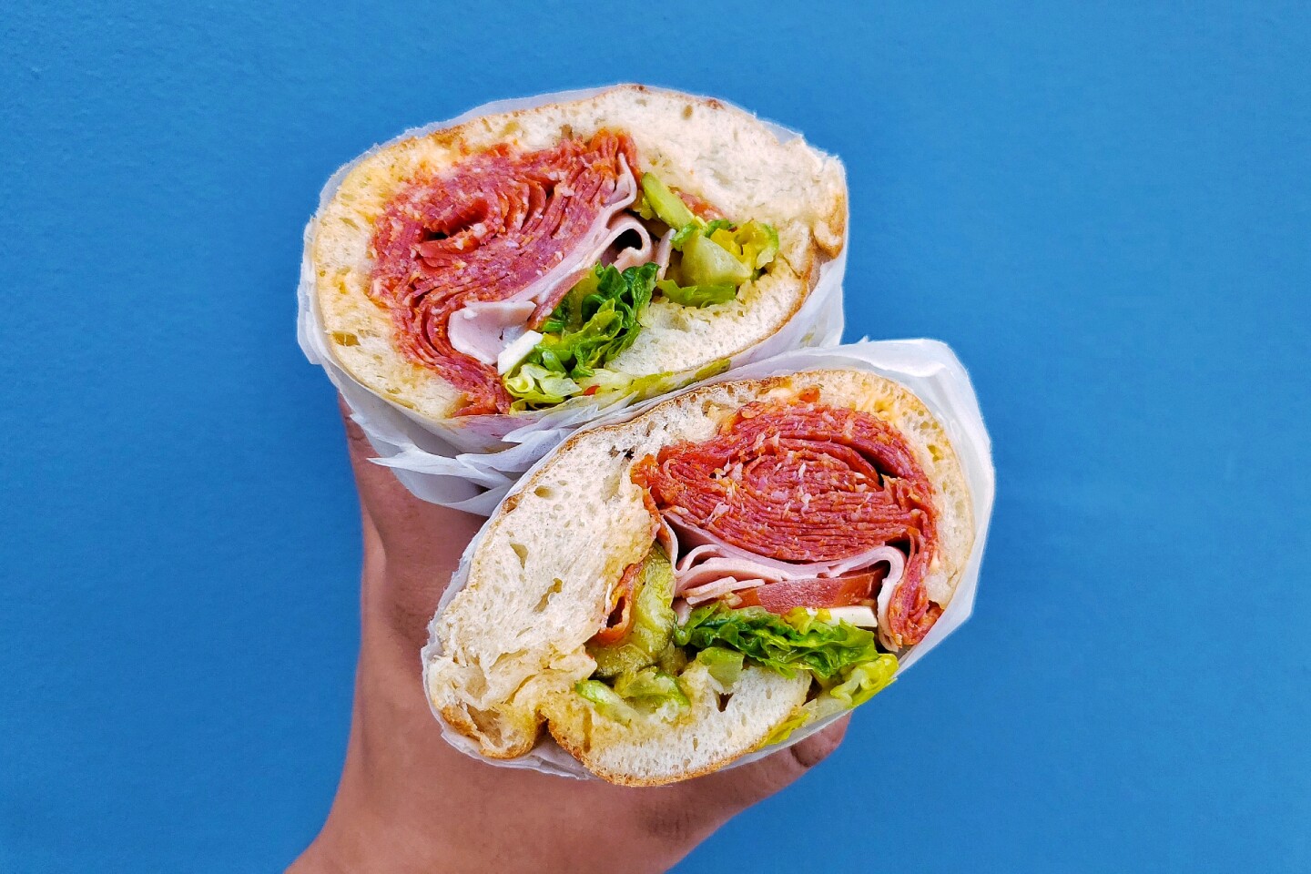 A hand holds two halves of a sandwich of sliced lunch meats, peppers and greens against a blue wall.
