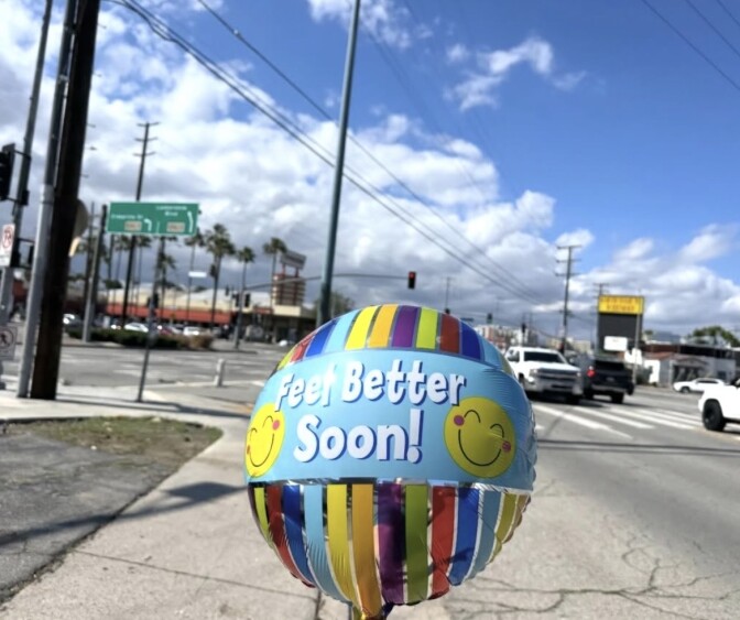 A small colorful balloon with yellow smiley faces and "feel better soon!" in blue-white text on the front is floating above a cement sidewalk near an intersection in the Los Angeles area.