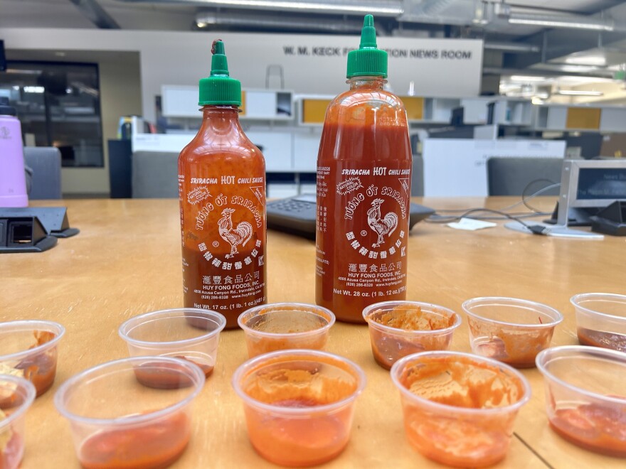 A smaller, mostly empty bottle of Sriracha and a larger, mostly full bottle sit behind two rows of clear used condiment cups.