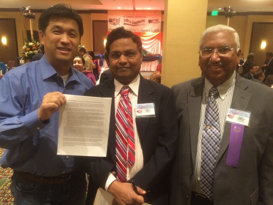 A man of East Asian descent (far left) holds a piece of printed paper while standing next to two men of South Asian descent who are wearing eyeglasses and suits. 