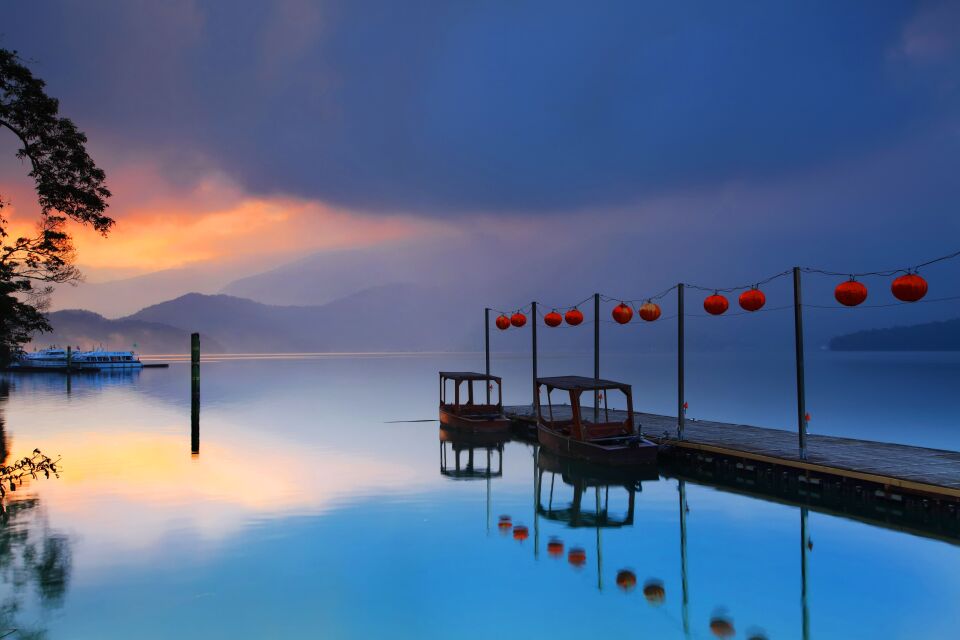 Small boats docked to a pear, red lanterns hang from pool running along the pear. The sun sets in the distance. 
