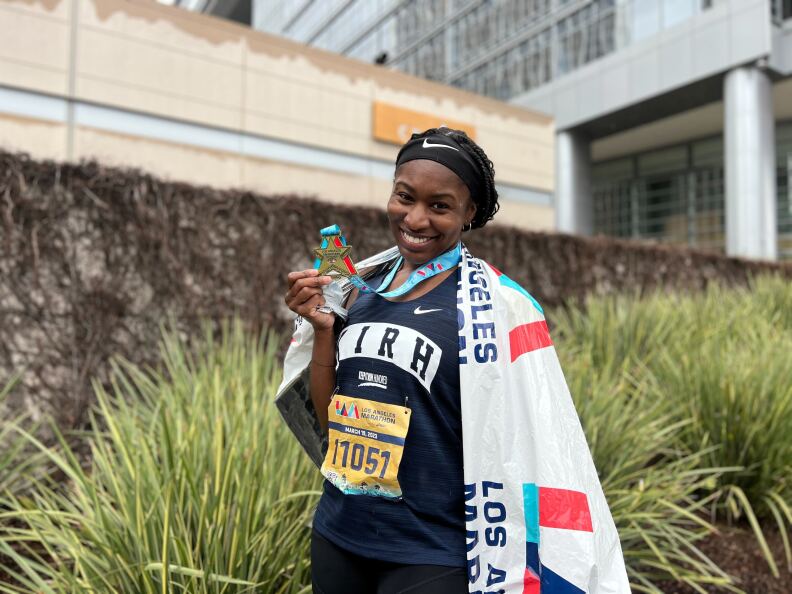A Black woman stands holding a medal, wearing a dark running shirt with a number pinned on the front. She is draped with a white wrapper post L.A. Marathon
