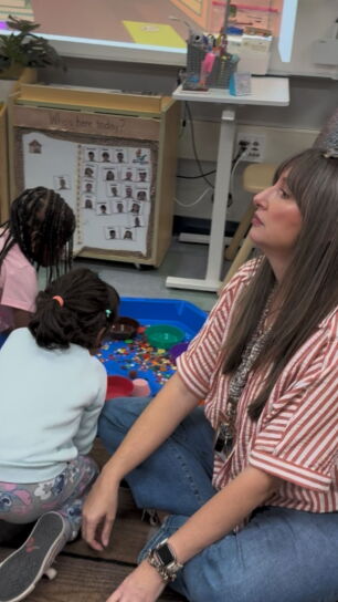 A preschool teacher sits on the floor with two students.