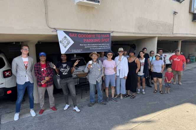 A group of people stand in front of low-level parking lots under an beige apartment complex. A sign hangs above one parkign spot that says, "Say goodbye to your parking spot." 