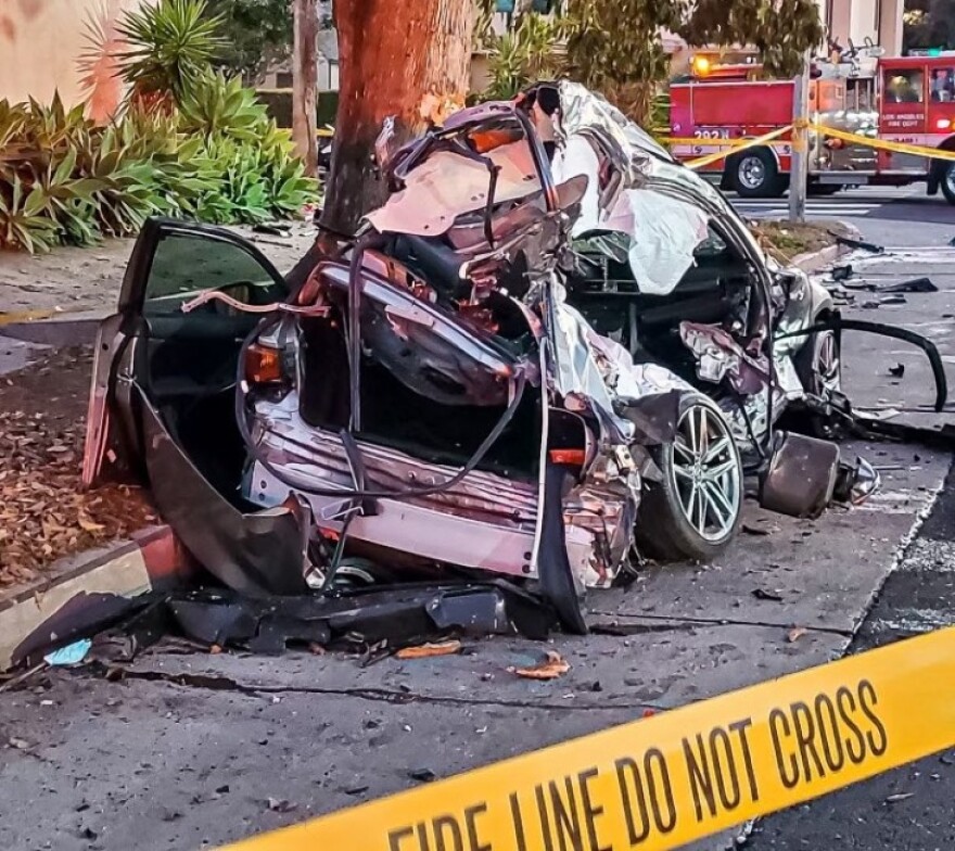 The crumpled remains of a car following a high-speed collision is behind police tape. The vehicle is mangled and misshapen.