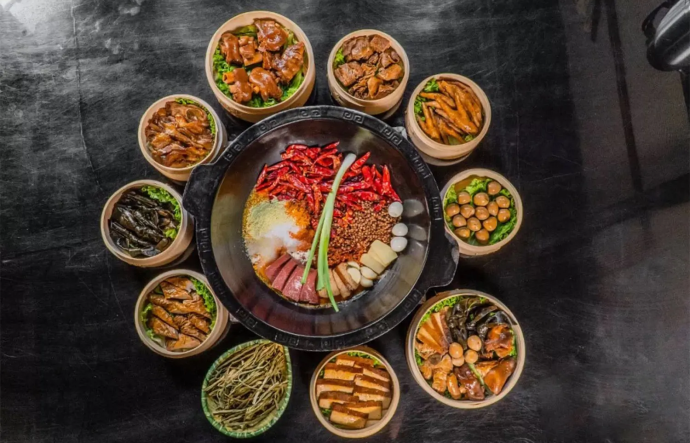 A large grey-colored stone dish containing a variety of meats and vegetables sits in the center of an assortment of round, light brown-colored steamer baskets, each containing different types of food on a black surface. 