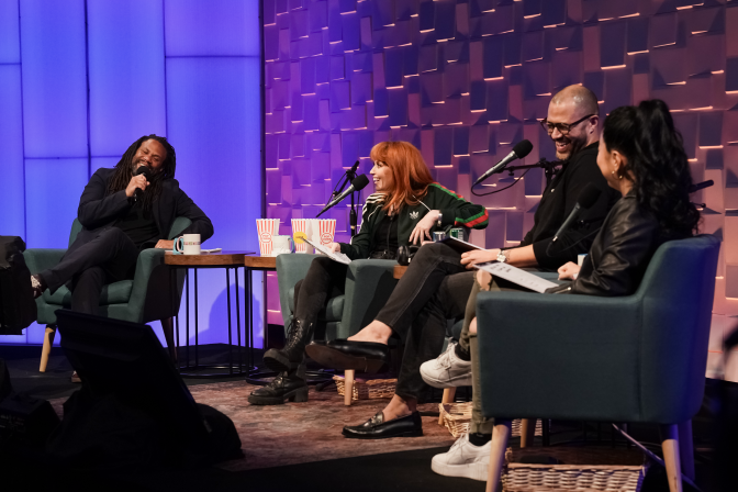 Four people are on a stage, sitting in green upholstered chairs with microphones in front of them. The person on the far left is a Black man wearing a black suit and shirt. To his left are 3 people: A red-headed white woman wearing a green and red track suit jacket and black pants; a biracial man with a shaved head and glasses; an Asian woman with long black hair in a ponytail. The last woman's face isn't visible. She is facing the other guests and host.