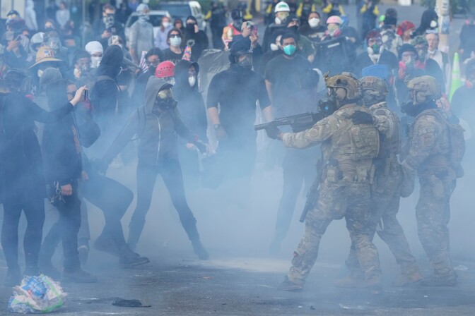 Federal agents in gear wearing masks hold riot guns pointing them directly to a group of protestors, who are also wearing masks. A hazy gas fills the area outside.