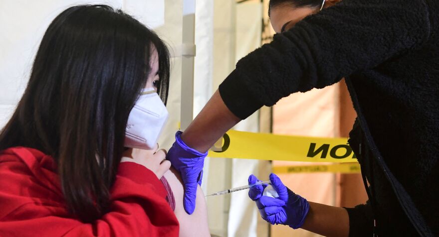 Kelly Zhou, 17, wears a white mask and holds the sleeve of her red shirt sleeve up as a gloved health care work gives her the Pfizer shot in her upper left arm.