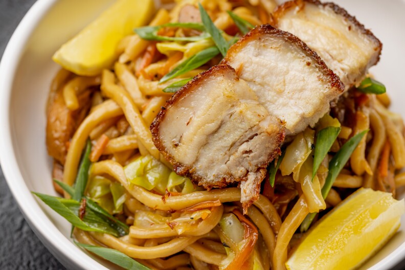 A white bowl of thick cooked noodles topped with three pieces of fried pork belly, thin cuts of green onion, and two slices of lemon.