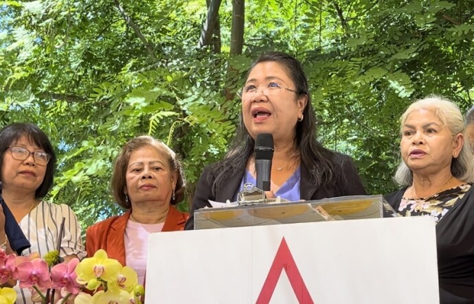 A 65-year-old Thai woman with long dark hair and glasses stands at a podium outside while three other women of Asian descent flank her. 