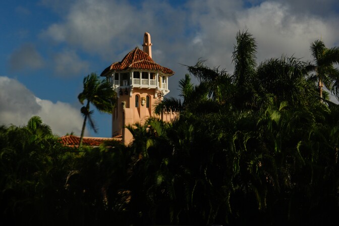 Mediterranean-style tower with terracotta-tiled roof and white balcony, partially obscured by lush palm trees against a blue sky with white clouds.