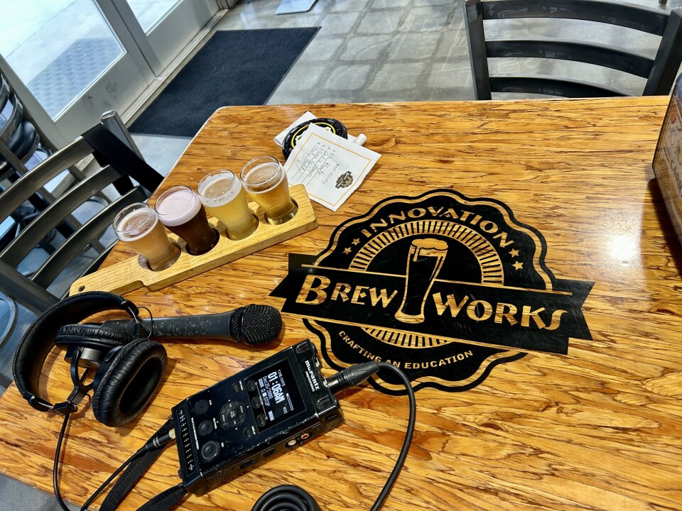 Four clear glasses with liquid rest on a wood table. A microphone and an audio recorder are next to the glasses.