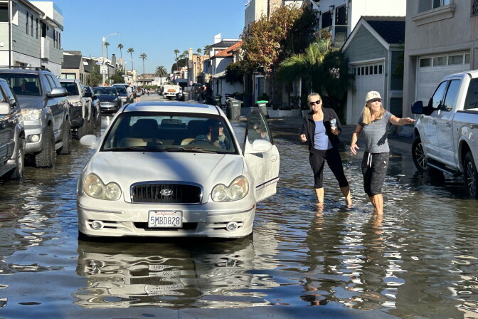 A car is stopped on a flooded residential street, where two women are wading through shin-deep water.