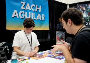A young man with brown hair wearing a white t-shirt and black lanyard looks down to sign an illustration. A man with brown hair and a black t-shirt stands across the table from him. Behind the man with the white shirt is a black curtain wall and blue and yellow banner that reads "Zach Aguilar" in white block letters.