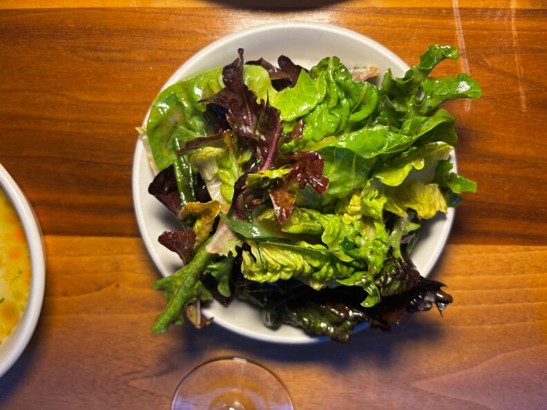 A simple mixed green salad served on a white plate, featuring a variety of fresh, leafy greens including romaine, red oak, and butter lettuce, lightly dressed and glistening under warm lighting on a wooden table.