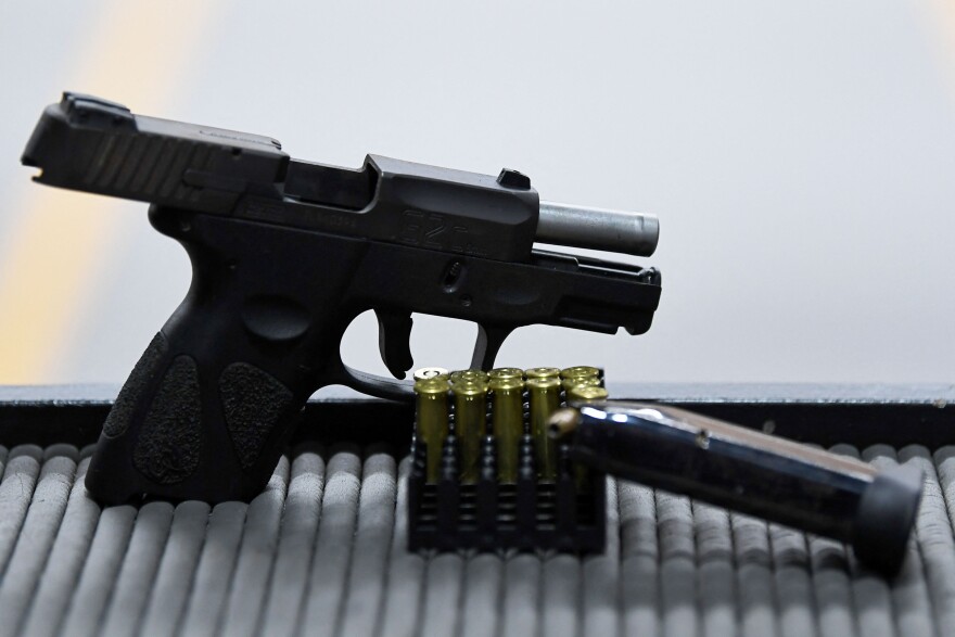 A black handgun sits with its magazine discharged on a table in front of an obscured gun range hall. A stack of gold-colored bullets sits in a case in between the gun and the gun's black magazine. The table is light gray with tubed texturing.