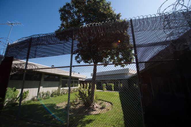 Looking through a mesh metal fence with razor wire on top. On the other side, people in neon green jail clothes are walking and standing outside of a building. 