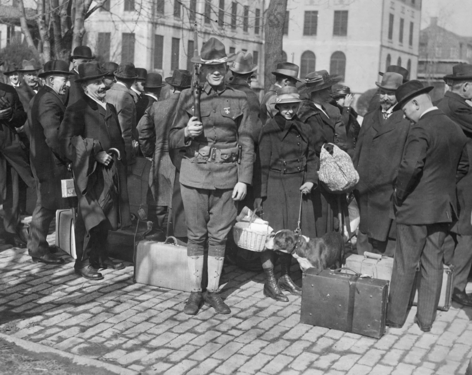 A black and white photo of many people with suitcases/briefcases and hats. There is a many that stands squarely for the camera in the center.