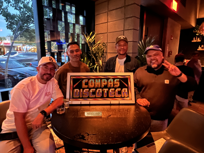 Four men pose at a table around a sign that says "Compás Discoteca". The one on the far left wears a white L.A. Dodgers hat. The one to the right of him wears a brown shirt. The one to the right of him wears a hat, blue jacket, and white shirt. The one one on the far right wears a blue hat and a Carhartt jacket.