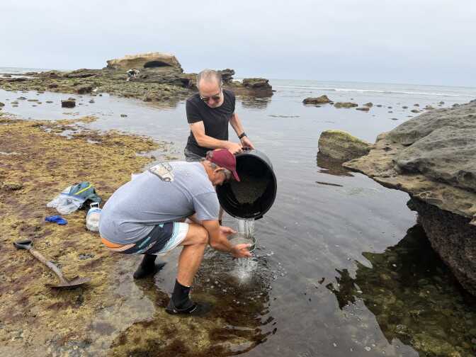 A man in a gray shirt squats in a tide pool while another man wearing black pours water from a large pail. The ocean is calm and the skies are overcast. 