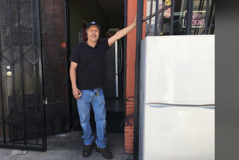 A man in a black shirt and blue jeans leans against a white refrigerator outside a storefront.