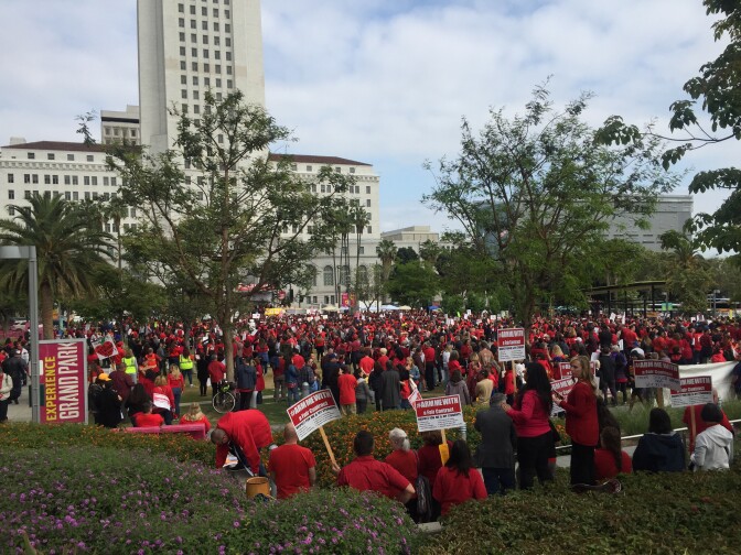 Teachers in bright red shirts holding signs and ringing red bells file into Grand Park. Representatives for the union say they chose to escalate their actions to a public rally after about a year of working without a contract. 