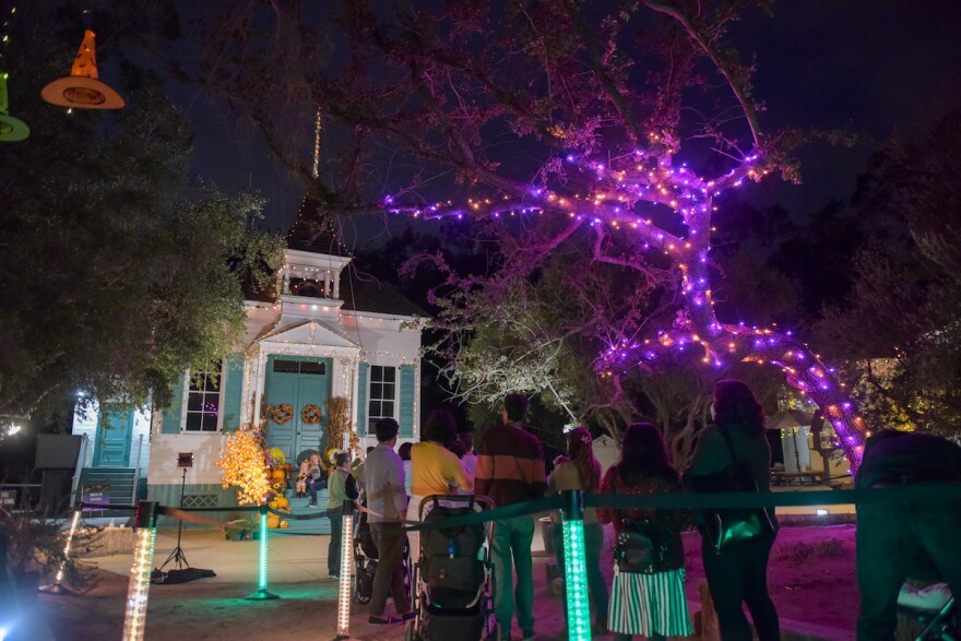 An old house decorated for Halloween with purple and orange lights with people milling out front. 