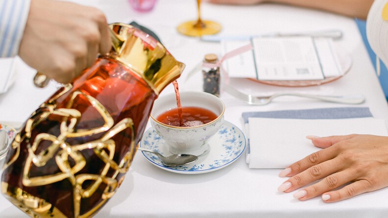 A person is pouring tea from a decorative red and gold teapot into a floral-patterned teacup on a white tablecloth. The table is set with various items, including a folded napkin, utensils, and another person's hand resting near the cup.