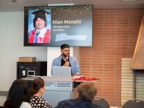 A man with medium brown skin, short dark hair, and a beard stands at a lectern in front of a classroom. He's holding a microphone with his laptop open in front of him. Behind him is a screen with a picture of a smiling person with a graduation cap and the words "CSU Bakersfield."