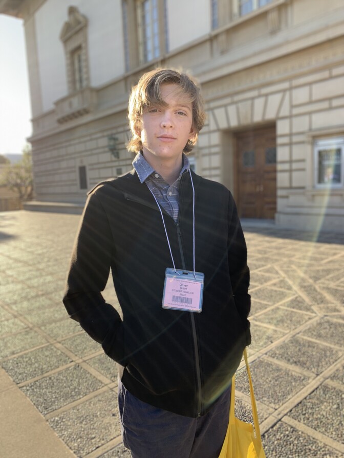 Young man with light skin and dark jacket stands in front of building with stone facade.