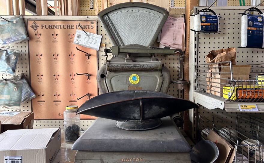 A close-up of a dusty metal scale sitting on top of a store shelf. The scale has a "DAYTON" logo in aged yellow towards the bottom, with a bright yellow and blue sticker in the middle. The scale is surrounded on all sides by metal hooks, merchandise, and screws. 