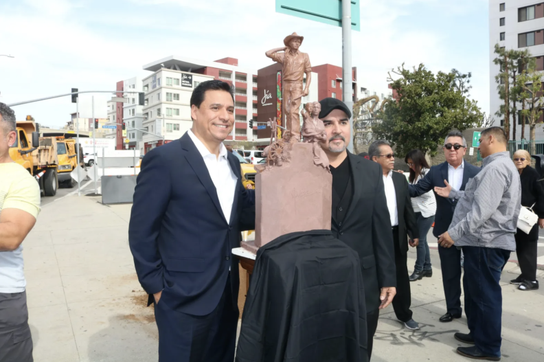 Two brown-skinned men pose of a picture, standing on each side of a small statue.