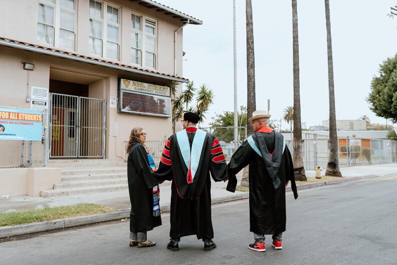 Holding hands and clad in graduation regalia, three people stand before a gated school. There are several palm trees in the background.
