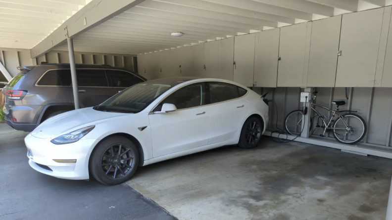 A white Tesla sits in a parking garage, charger attached to the wall.