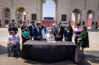 A group of people, including several in USA Olympic and Paralympic team apparel, gather around a table at the entrance arch of the Los Angeles Memorial Coliseum for a document-signing ceremony. A seated official signs papers at the black-draped table while others look on and smile, with the stadium's stands and bronze plaques visible in the background.