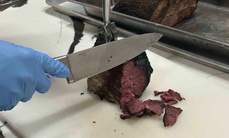 a slab of pink pastrami is being cut with a large knife by a hand in a blue glove