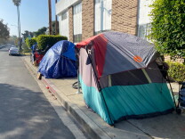 Three tents used by unhoused people are shown on a sidewalk next to an apartment building.