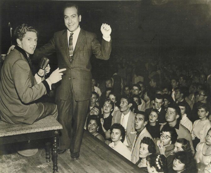A man in a dark suit raises his left arm on stage, talking to a performer. There's a big crowd in the audience.